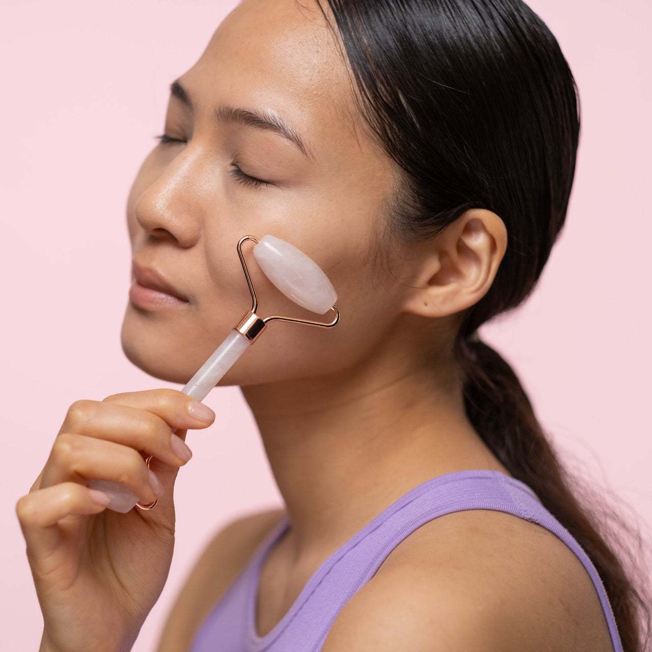 Young woman using a jade roller for her skin care routine on a pink background.